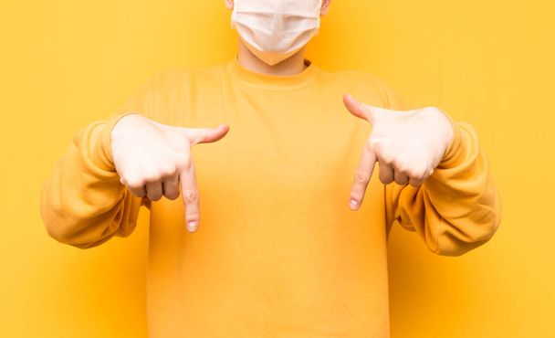 Close photo. Background. Person in the medical mask points his finger down at the copy space. Coronavirus pandemic. Quarantine. covid-19. Man in a protective mask and bright clothes points down. - Φωτογραφία, εικόνα