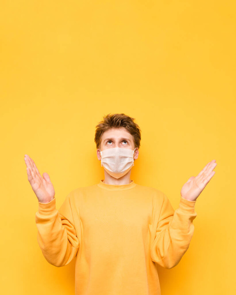 Σοβαρός τύπος με πορτοκαλί μπλούζα και ιατρική μάσκα στέκεται σε κίτρινο φόντο, απλώνει τα χέρια του στα πλαϊνά και κοιτάζει το χώρο των αντιγράφων. Πανδημία του κορωναϊού. Καραντίνα. κοβίδιο-19. - Φωτογραφία, εικόνα