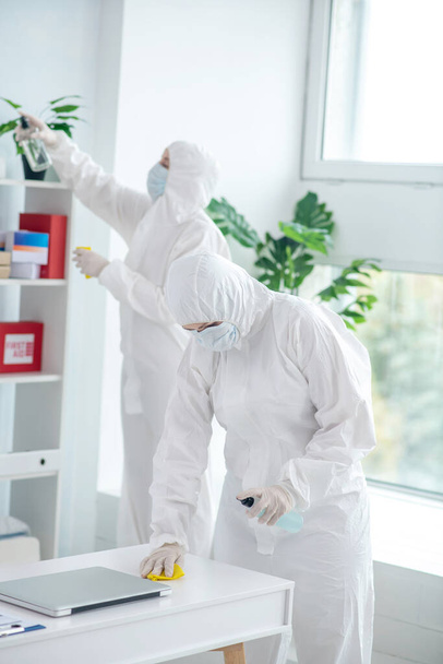 Medical worker in protective clothing and medical mask disinfecting table, another worker cleaning closet - Photo, image
