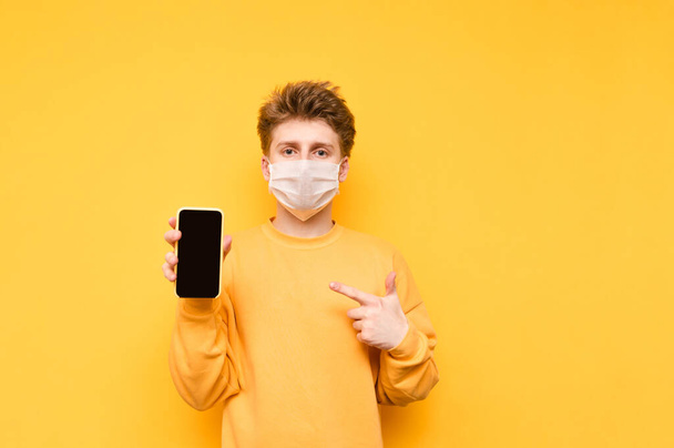 Handsome guy in a gauze mask stands with a smartphone in his hand on a yellow background, points his finger at the black screen and looks at the camera with a serious face. - Fotoğraf, Görsel
