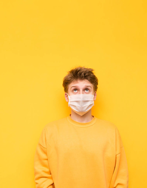 Portrét zamyšleného mladíka ve žlutém svetru a bílé lékařské masce izolované na žlutém pozadí, hledícího na kopírovací prostor a přemýšlejícího. Koronavirová pandemie. Karanténa. covid-19. - Fotografie, Obrázek