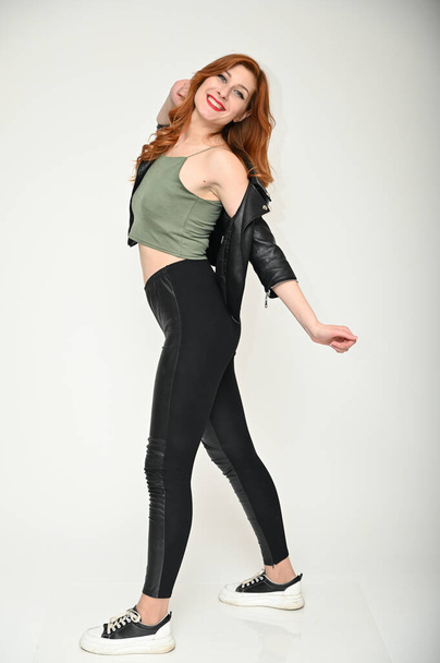 Full-length photo of a young Caucasian woman with excellent makeup with long red hair in a black jacket and trousers. Caucasian model poses with different emotions on a white background in the studio. - Фото, зображення