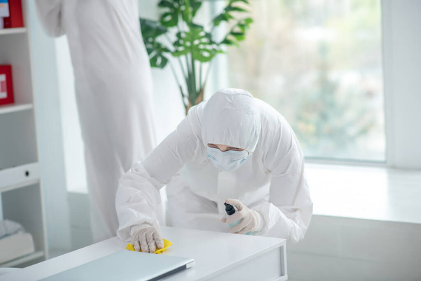 Medical worker in protective clothing and medical mask disinfecting table, another worker standing behind - Φωτογραφία, εικόνα