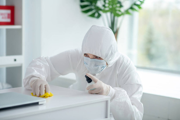Medical worker in protective clothing and medical mask sprays disinfectant on table - Φωτογραφία, εικόνα