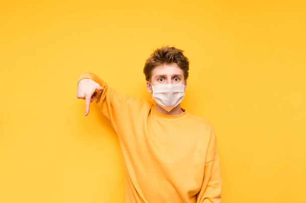 Emotional young man in bright clothes points his finger down at the copy space and looks at the camera with a shocked face, isolated on an orange background. Coronavirus pandemic. Quarantine. covid-19 - Photo, Image