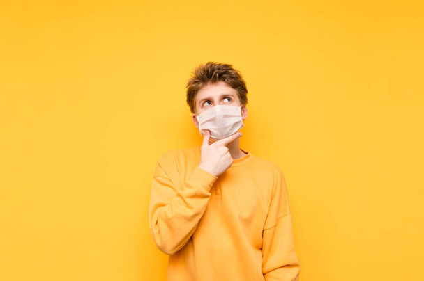 Πορτρέτο ενός σκεπτικού τύπου με ιατρική μάσκα σε κίτρινο φόντο, κοιτάζοντας το χώρο αντιγραφής και τη σκέψη. Έξυπνος τύπος με προστατευτική μάσκα σκέφτεται τον κορωναϊό. Πανδημία. Καραντίνα. κοβίδιο-19. - Φωτογραφία, εικόνα