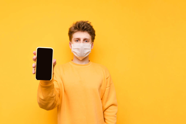 Knappe vent in een geel sweatshirt en een beschermend gaasmasker staat op een oranje achtergrond en toont de camera een smartphone met een zwart scherm. Coronavirus pandemie. Quarantaine. covid-19. - Foto, afbeelding