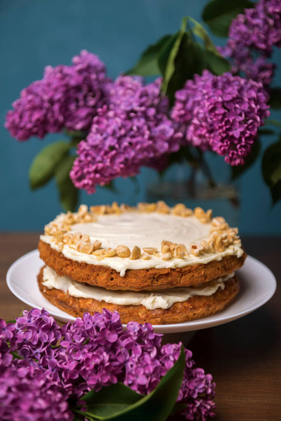 Carrot cake with hazel nuts and bouquet of purple lilac on the wooden table, pie cooked with carrot biscuit and sour cream, decorated with hazel nuts - Photo, Image
