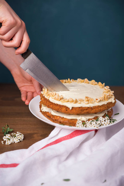 Руки режут морковный торт с орехами ореха, украшенный белыми цветами луговой сладости на деревянном столе
 - Фото, изображение