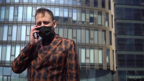 A young guy in a business suit and a black mask talking on the phone in the background of an office building. - Video