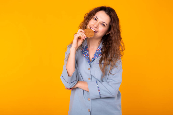Radostná žena nosí módní pyžama bude jíst lahodné sušenky na žlutém pozadí s kopírovacím prostorem. Ranní, snídaně a domácí opotřebení koncepce. - Fotografie, Obrázek