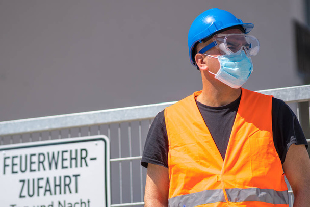 Sicurezza e protezione del lavoro contro le condizioni avverse sul lavoro. Operaio edile con cappello rigido blu, gilet riflettente e maschera protettiva chirurgica
 - Foto, immagini