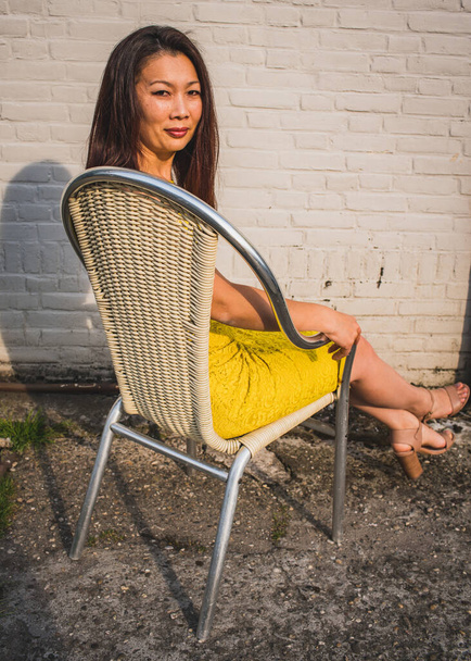 Азиатская модель среднего возраста в желтом платье сидит на фоне белой кирпичной стены, на открытом воздухе, Промышленное пространство
. - Фото, изображение