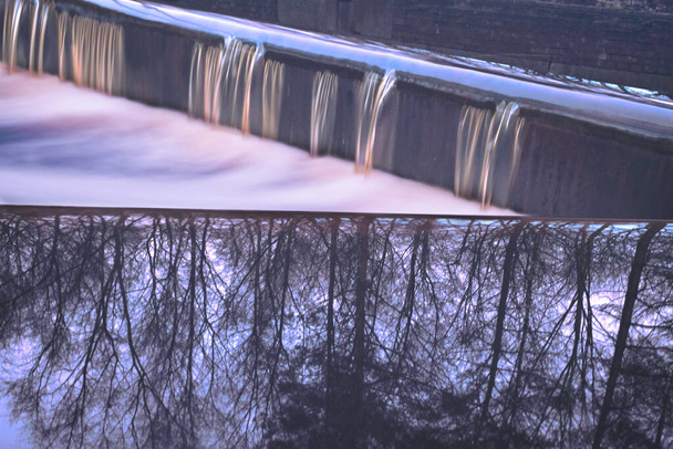 En un vertedero fluye mucha agua a lo largo del vertedero, reflejo de árboles en el agua, movimiento del agua con exposición prolongada. Ubicación: Frisia, Países Bajos
. - Foto, Imagen