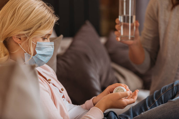 Selektiver Fokus des Kindes in medizinischer Maske mit Pillen in der Nähe der Mutter mit einem Glas Wasser  - Foto, Bild