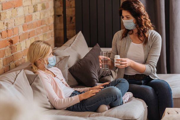 Selective focus of woman holding glass of water and jar with pills near child in medical mask on couch  - Photo, Image