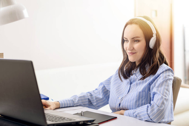 ευτυχισμένη καυκάσιος γυναίκα σε μπλε πουκάμισο με λευκά ακουστικά ακούγοντας μουσική, απομακρυσμένη εργασία στο σπίτι, ελεύθερος επαγγελματίας, άνετη ζωή - Φωτογραφία, εικόνα