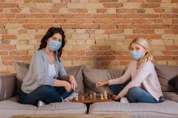 Vista lateral de la madre y la hija en máscaras médicas jugando ajedrez en el sofá
  - Foto, Imagen