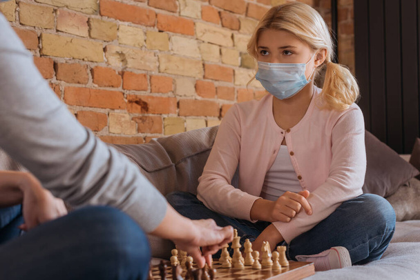 Az orvosi maszkos gyerek szelektív fókusza, aki otthon sakkozik anyával  - Fotó, kép