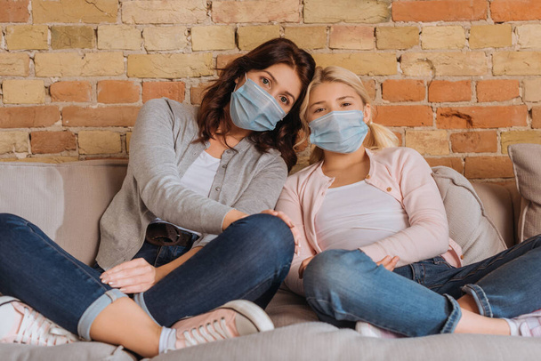 Mother and kid in medical masks looking at camera on couch at home  - Фото, зображення
