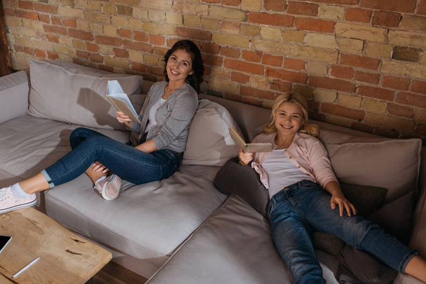 High angle view of smiling mother and daughter looking at camera while reading books on couch  - Foto, imagen