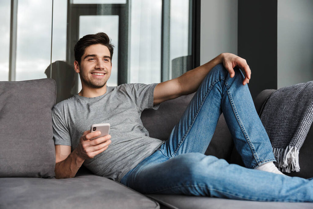 Portrait of an attractive smiling young bearded man wearing casual clothes sitting on a couch at the living room, using mobile phone - 写真・画像
