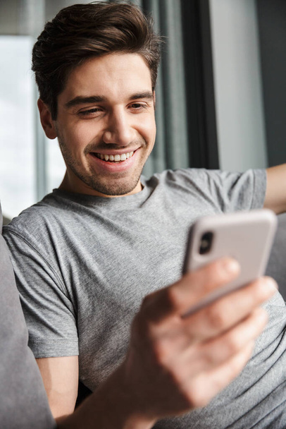 Portrait of an attractive smiling young bearded man wearing casual clothes sitting on a couch at the living room, using mobile phone - Фото, зображення