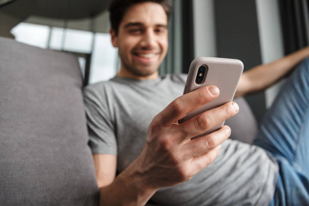 Portrait of an attractive smiling young bearded man wearing casual clothes sitting on a couch at the living room, using mobile phone - Fotó, kép