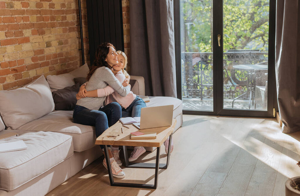 Smiling mother hugging child near laptop, notebook and book on coffee table at home  - Φωτογραφία, εικόνα