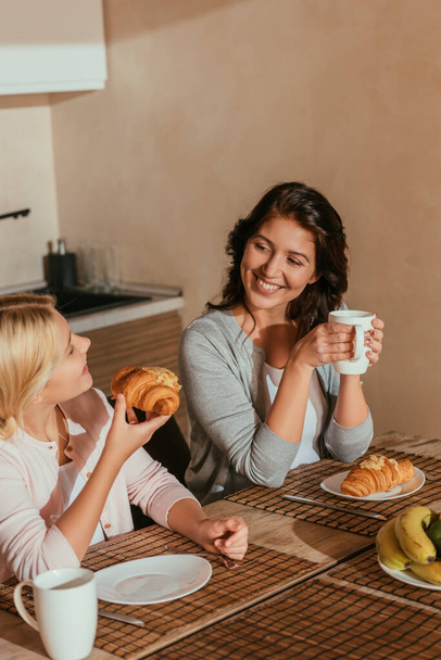 Selective focus of mother smiling at kid with croissant during breakfast in kitchen   - Foto, Bild