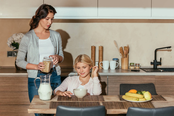 Smiling kid sitting at table near mother holding jar with cereals in kitchen  - Foto, Imagen