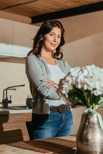 Selective focus of beautiful woman with crossed arms looking at camera near flowers on kitchen table  - Foto, immagini