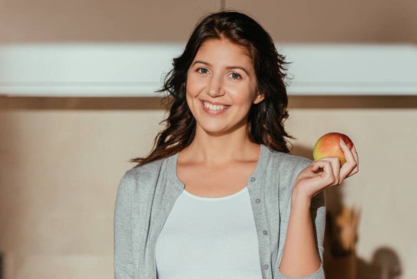 キッチンで新鮮なリンゴを保持しながら、カメラを見て美しい笑顔の女性  - 写真・画像
