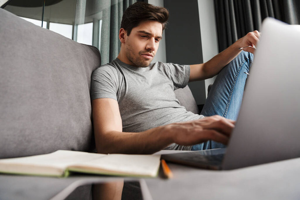 Portrait of an attractive serious young bearded man wearing casual clothes sitting on a couch at the living room, using laptop computer - Φωτογραφία, εικόνα