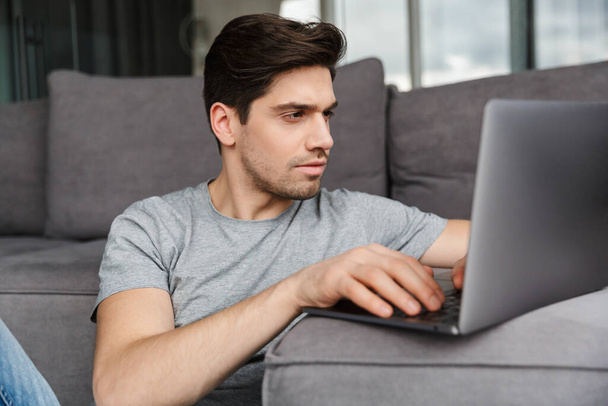 Portrait of an attractive serious young bearded man wearing casual clothes sitting on a floor at the couch in the living room, using laptop computer - Foto, afbeelding