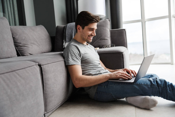 Portrait of an attractive smiling young bearded man wearing casual clothes sitting on a floor at the couch in the living room, using laptop computer - Zdjęcie, obraz