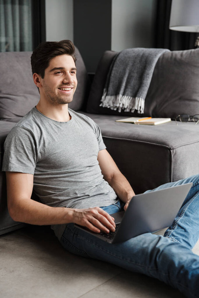 Portrait of an attractive smiling young bearded man wearing casual clothes sitting on a floor at the couch in the living room, using laptop computer - Valokuva, kuva