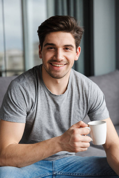 Image of a happy handsome young man indoors at home drinking coffee. - Fotoğraf, Görsel