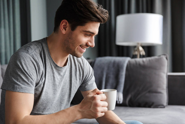 Image of a handsome cheerful young man indoors at home drinking coffee. - Foto, Imagem