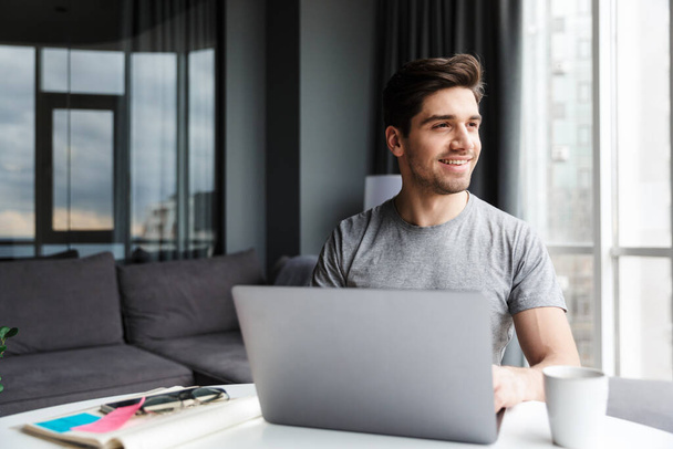 Handsome smiling young bearded man wearing casual clothes using laptop computer while sitting at the table at home - Foto, immagini