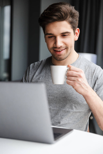 Bonito sorridente jovem barbudo vestindo roupas casuais usando computador portátil enquanto sentado na mesa em casa, bebendo café
 - Foto, Imagem