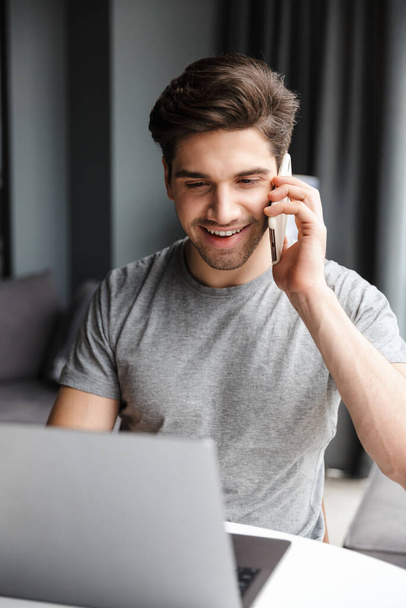 Bonito sorridente jovem barbudo vestindo roupas casuais usando computador portátil enquanto sentado na mesa em casa e falando no telefone celular
 - Foto, Imagem