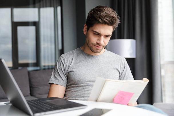 Image of a handsome young man indoors at home using laptop computer holding documents. - Foto, immagini