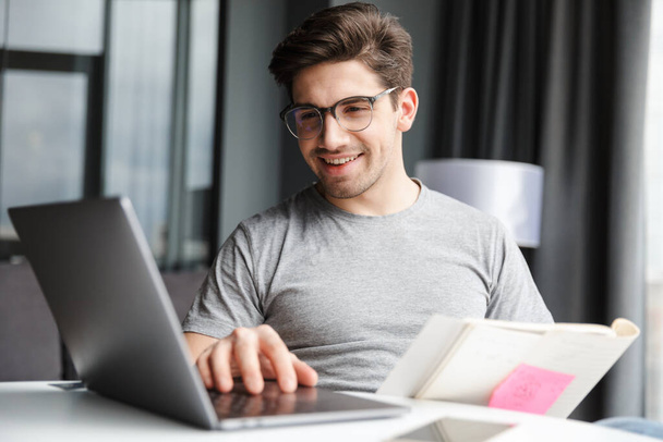 Image of a positive optimistic young man indoors at home using laptop computer holding documents. - Foto, Bild