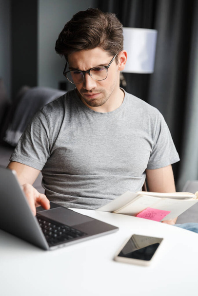 Handsome serious young bearded man wearing casual clothes using laptop computer while sitting at the table at home - Foto, Imagem