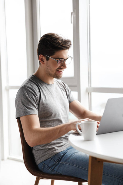 Handsome smiling young bearded man wearing casual clothes using laptop computer while sitting at the table at home - Фото, зображення