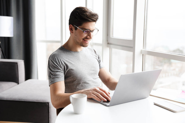 Handsome smiling young bearded man wearing casual clothes using laptop computer while sitting at the table at home - Foto, Bild