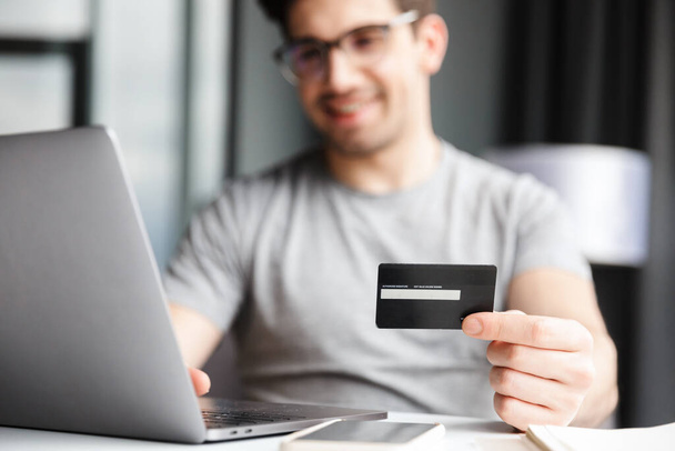 Handsome smiling young bearded man wearing casual clothes using laptop computer while sitting at the table at home, showing plastic credit card, shopping - Foto, immagini