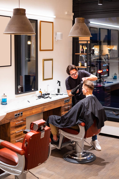 Homme coiffeur sèche les cheveux d'un jeune homme avec un sèche-cheveux et coiffe les cheveux avec une brosse dans son salon de coiffure
 - Photo, image