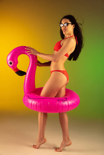 Fashion portrait of young fit and sportive woman in stylish red luxury swimwear with rubber flamingo on gradient background. Perfect body ready for summertime. - Zdjęcie, obraz